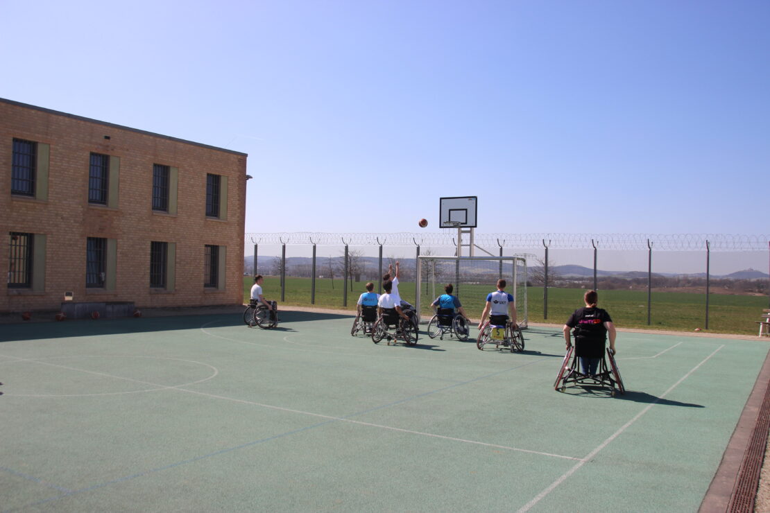 Mehrere Inhaftierte spielen Rollstuhlbasketball auf dem Sportplatz im Gefängnis