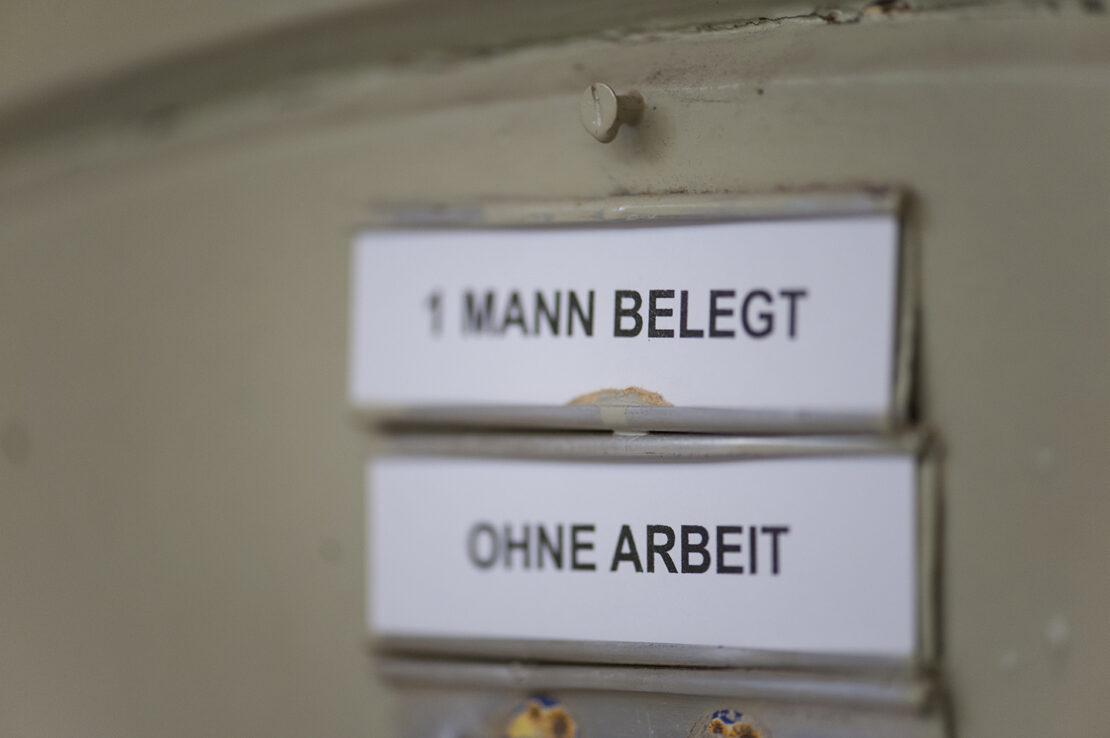 Zwei Schilder an Zellentür mit Aufschriften "1 Mann belegt" und "Ohne Arbeit"