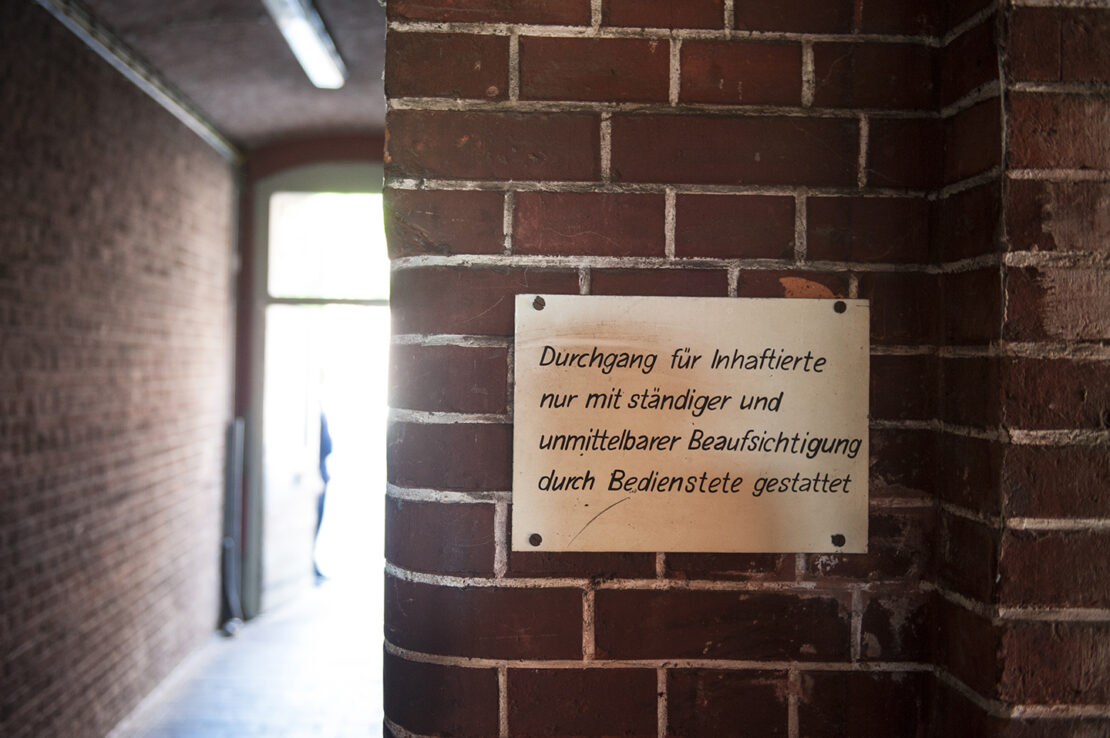 Dunkler Gang mit Schild an Wand, auf dem steht, dass Inhaftierte nur in Begleitung von Bediensteten hier langgehen dürfen